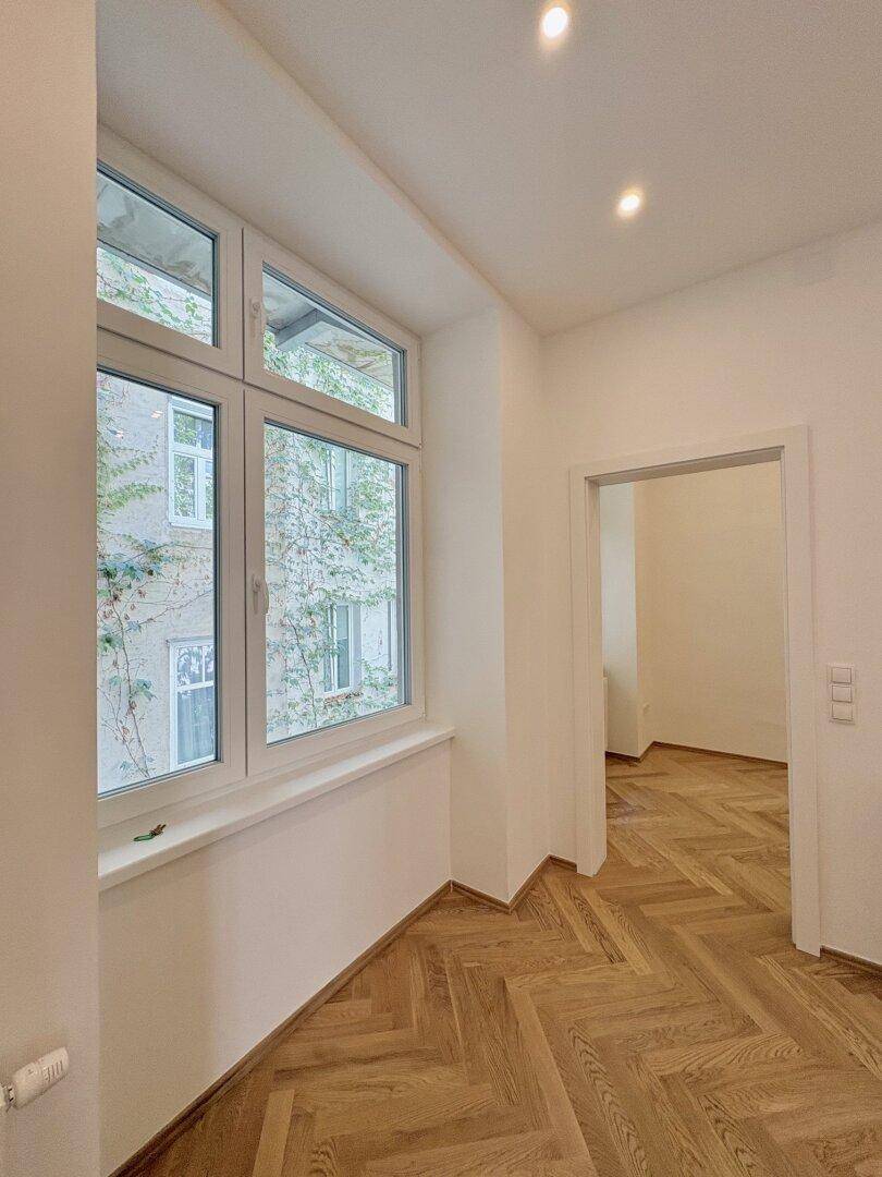 Heller Flur mit Fischgrätparkett und einem Fenster mit Blick auf den begrünten Innenhof.