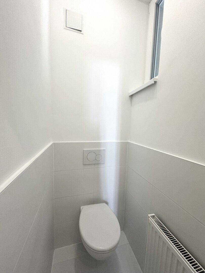 Separate Toilette mit wandmontiertem WC, kleinem Fenster und weißen Fliesen.