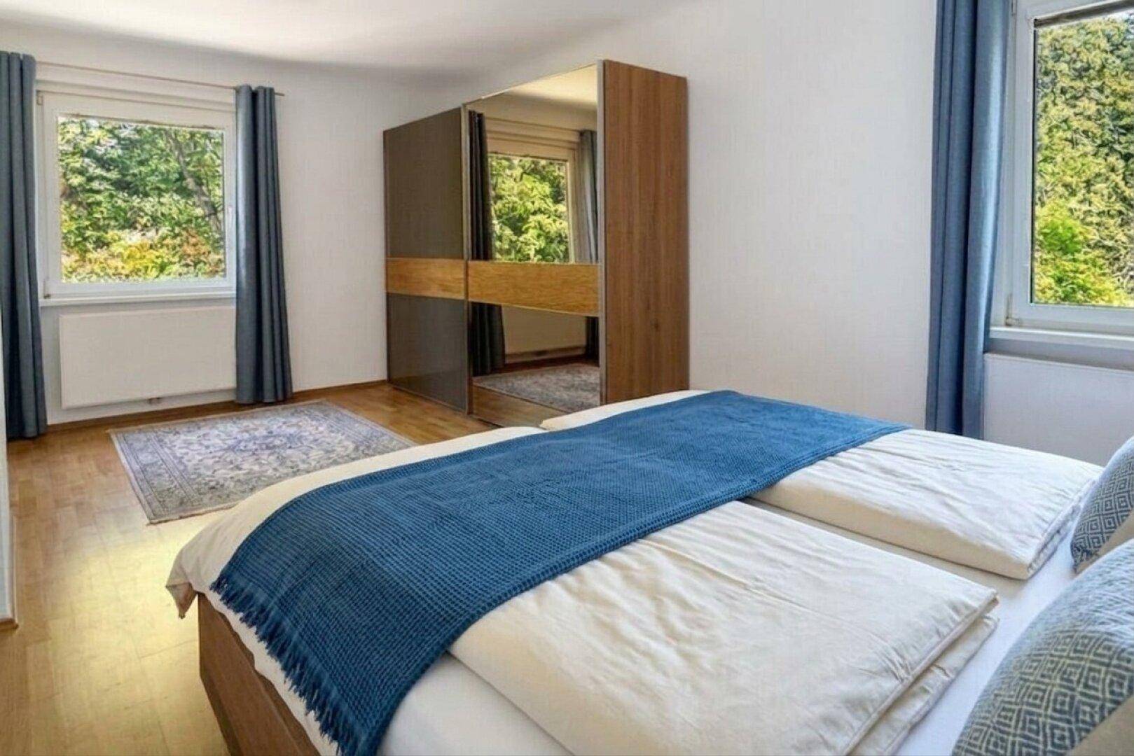 Schlafzimmer mit zwei Fenstern, Holzboden und einem großen Spiegelschrank.