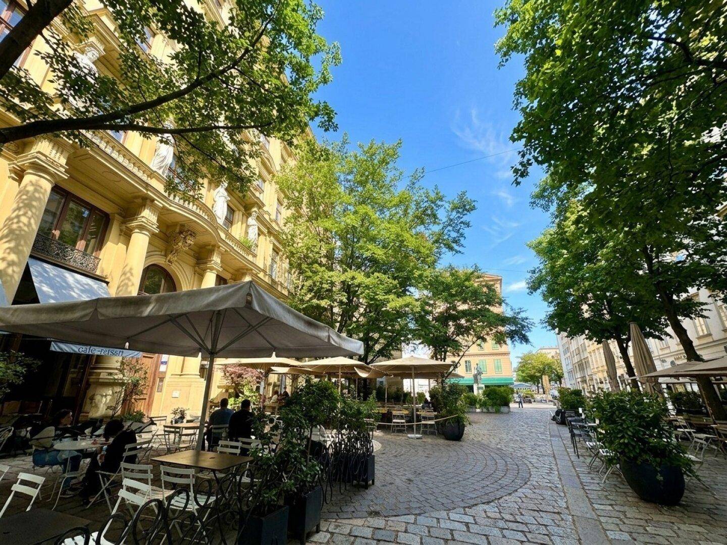 Belebte Kopfsteinpflasterstraße mit Straßencafés, historischen Gebäuden und schattenspendenden Bäumen an einem sonnigen Tag.