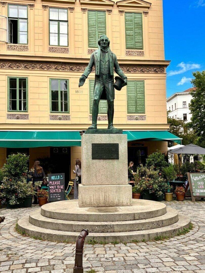 Historisches Gebäude mit grünen Fensterläden und einer Statue im Vordergrund auf einem belebten Platz.