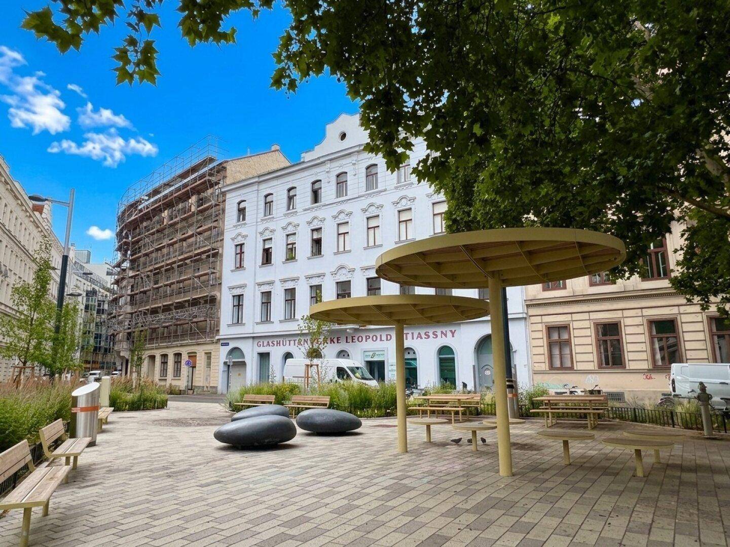 Moderner öffentlicher Platz mit Sitzbänken, Bäumen und einzigartigen Sonnenschutzstrukturen, umgeben von Gebäuden.