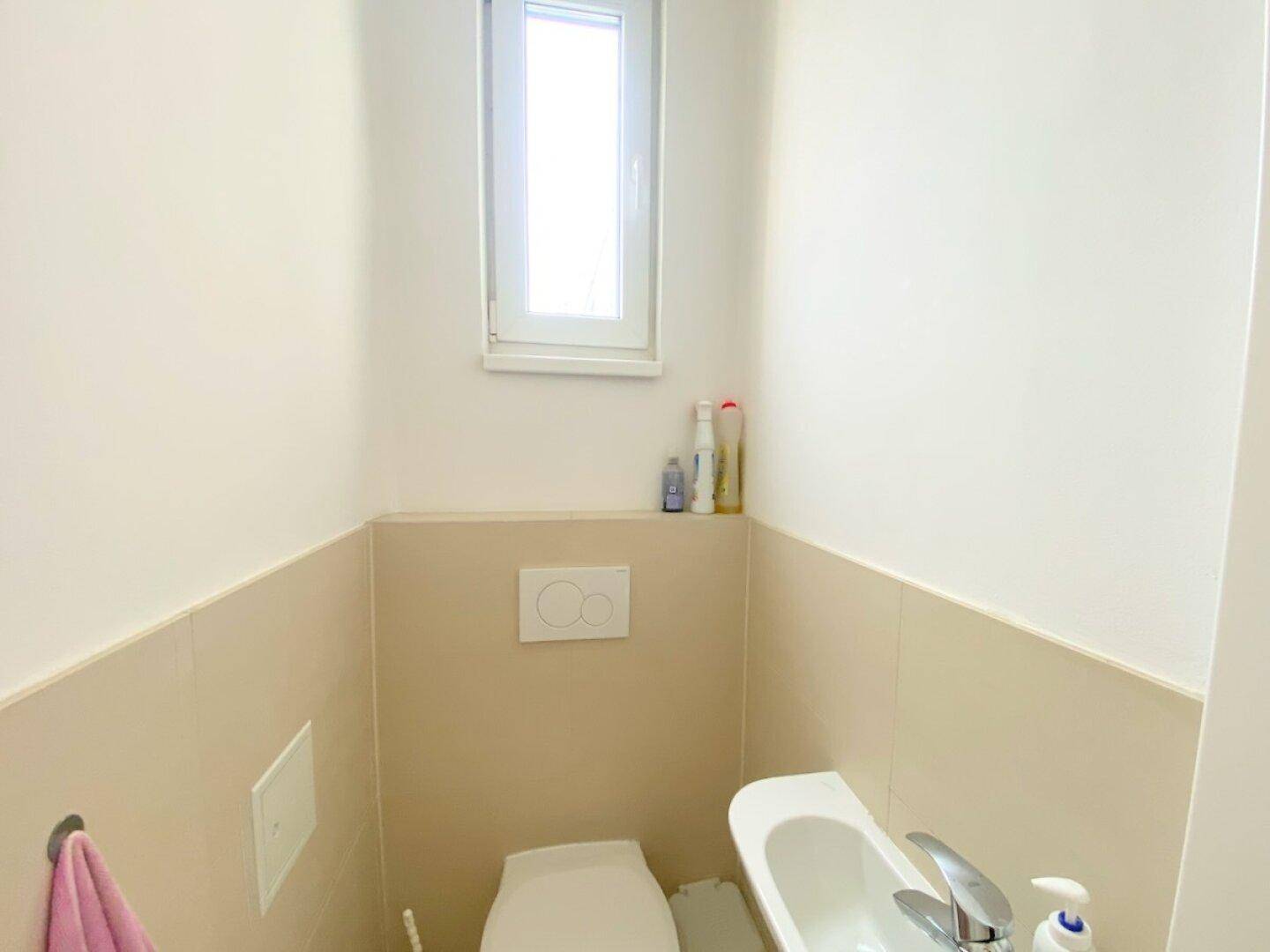 Kleines Gäste-WC mit Toilette, Waschbecken und Fenster für natürliche Belüftung, in hellen Farbtönen gehalten.