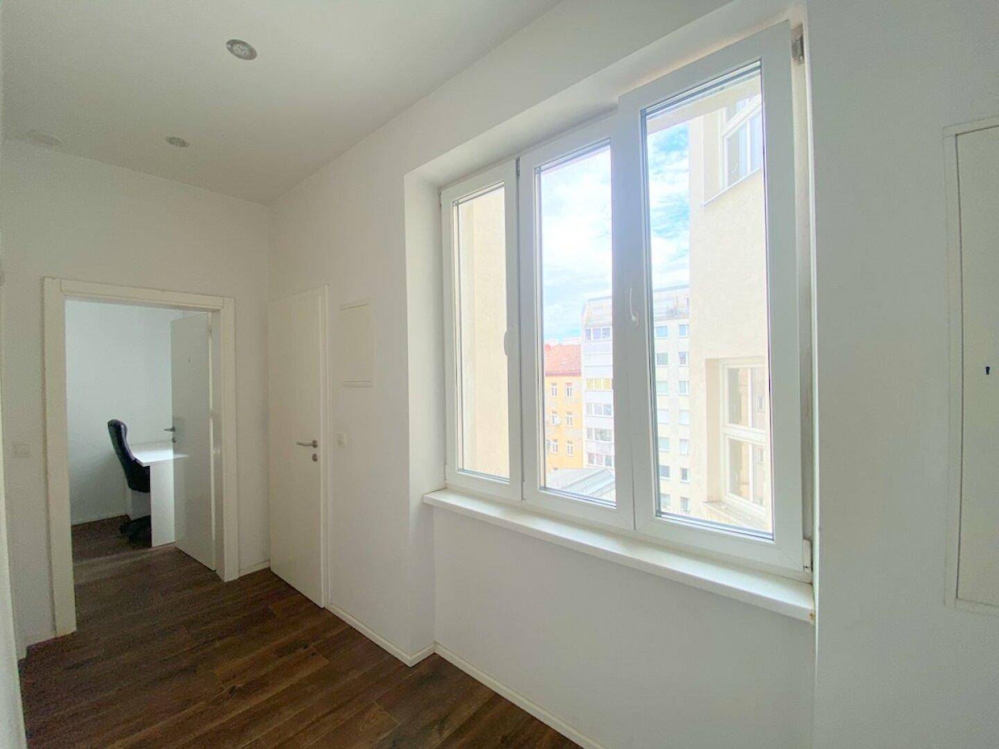 Heller Flur mit Holzfußboden und großem Fenster, das viel Tageslicht hereinlässt und zu weiteren Räumen führt.