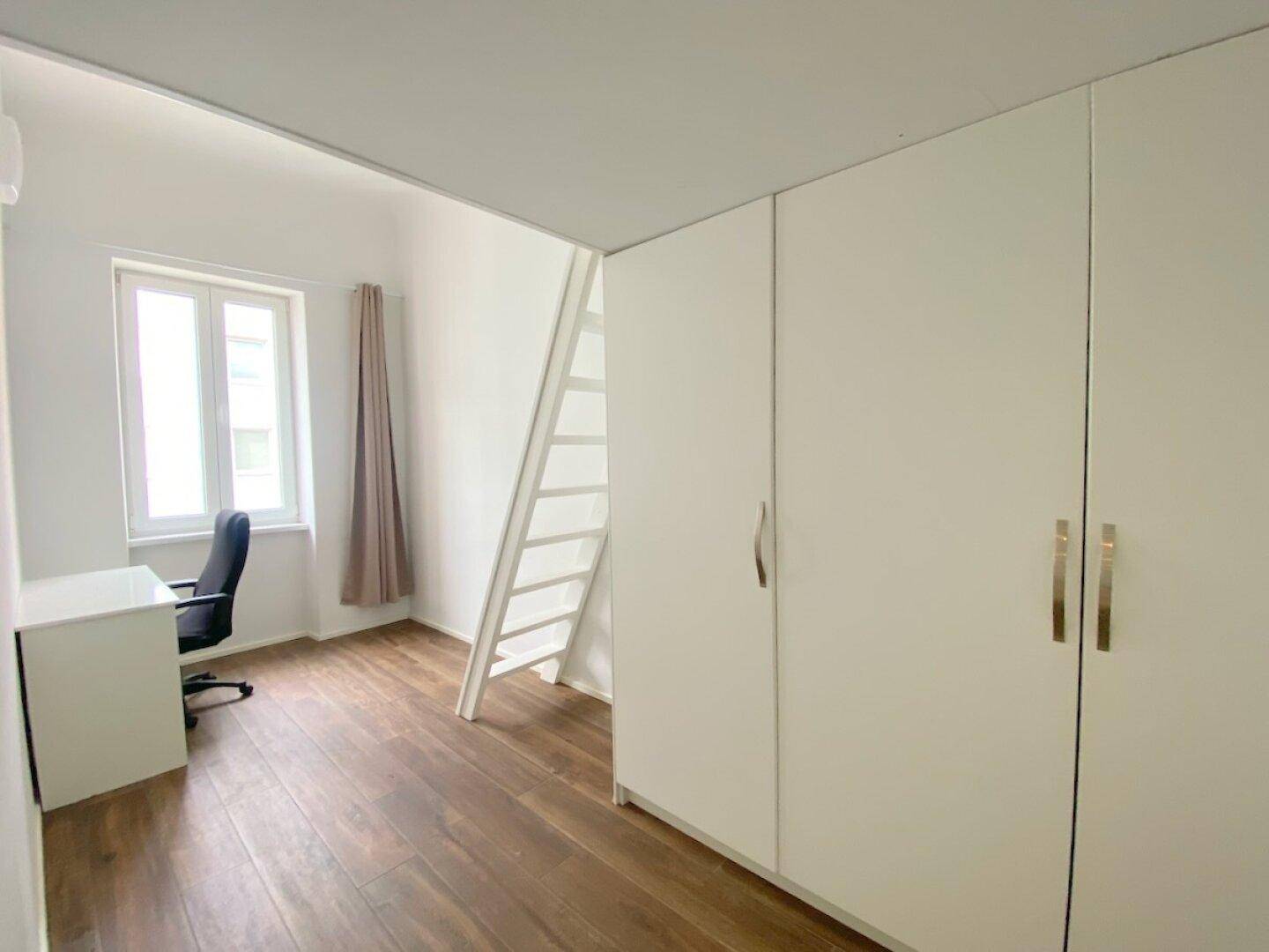 Geräumiges Zimmer mit Schreibtisch, Fenster und großem Kleiderschrank, ideal als Arbeitszimmer, auf Holzfußboden.