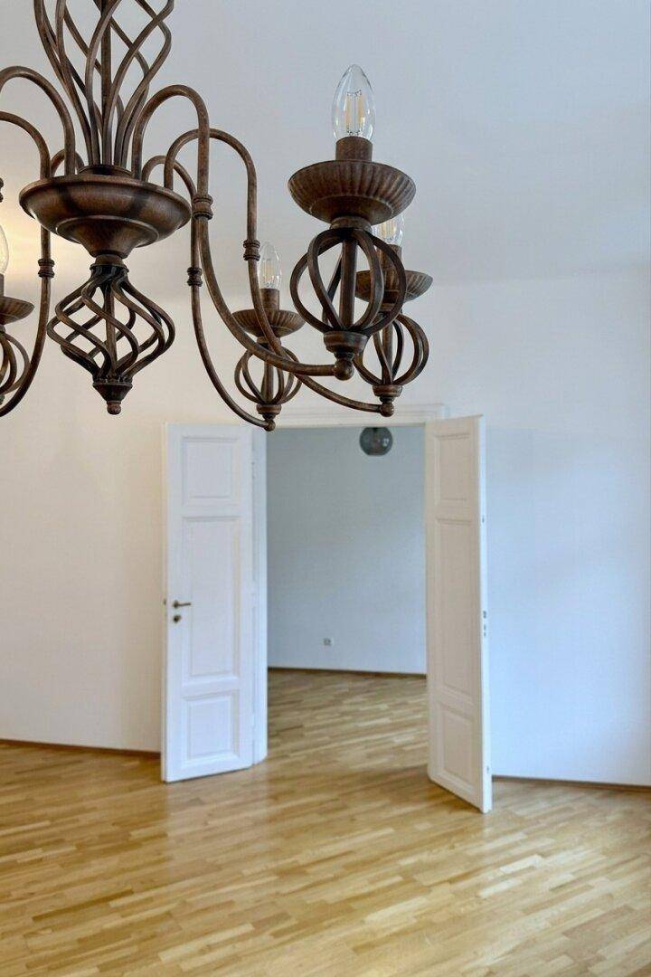 Detailaufnahme eines eleganten Kronleuchters über einem Raum mit Fischgrätparkett und Flügeltüren.