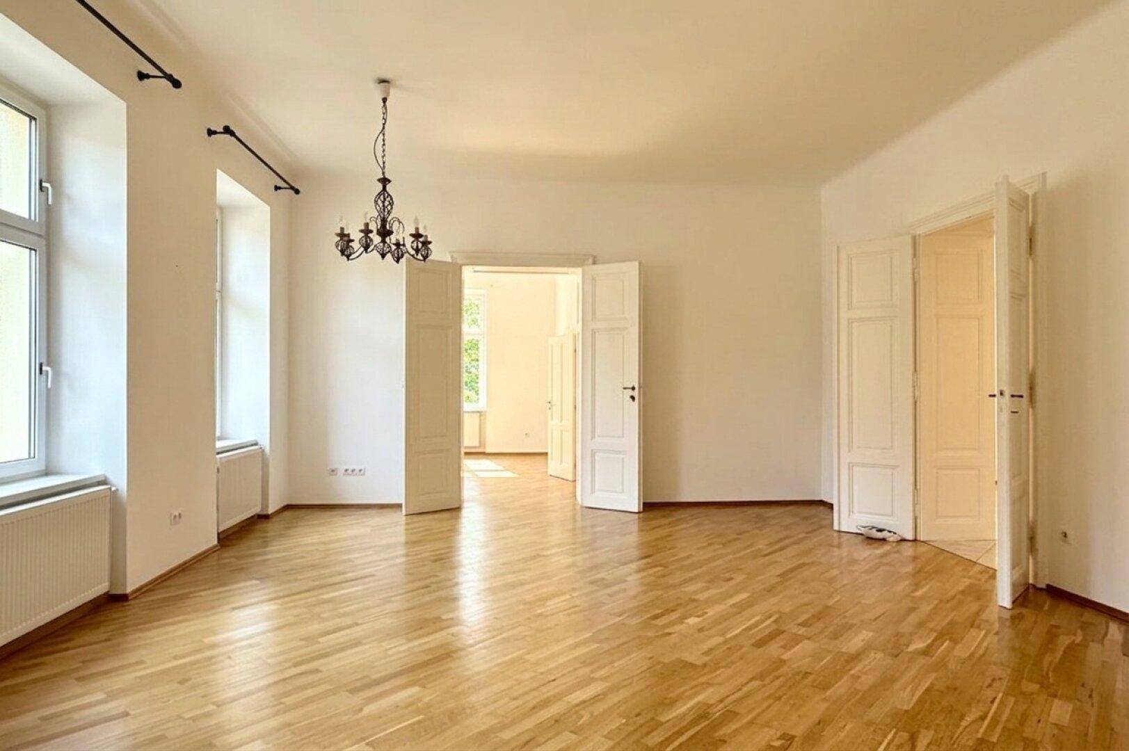 Großer, leerer Raum mit hellem Fischgrätparkett, hohen Fenstern und Zugang zu weiteren Zimmern.