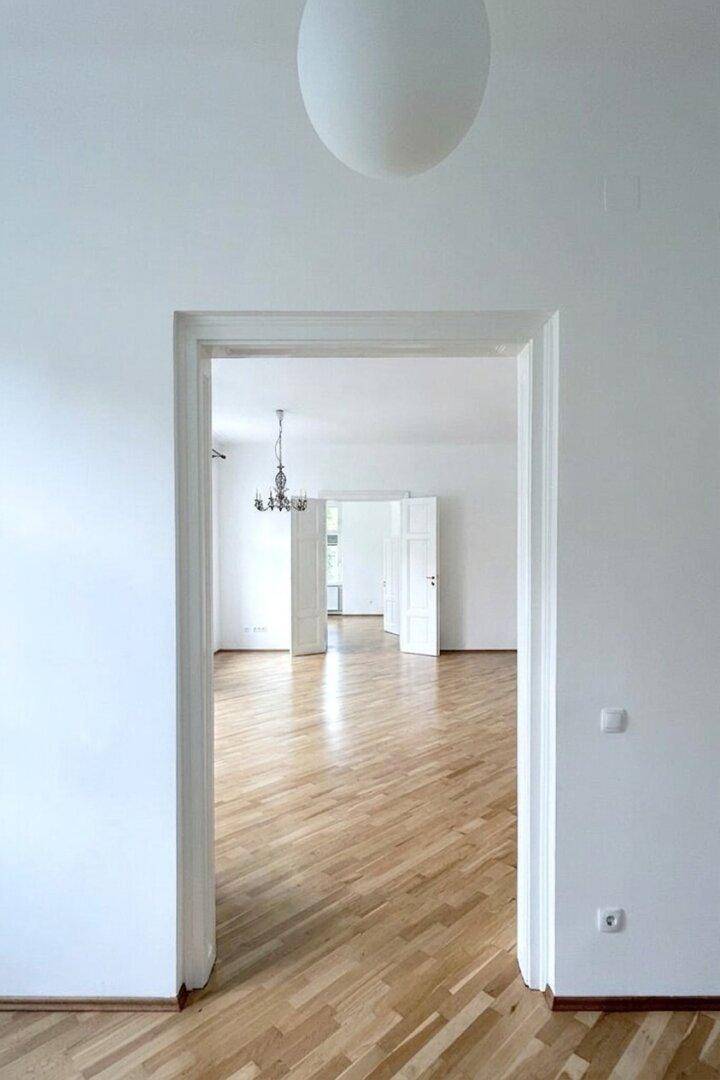 Blick durch eine Türöffnung in einen hellen Raum mit Fischgrätparkett und hohen Decken.