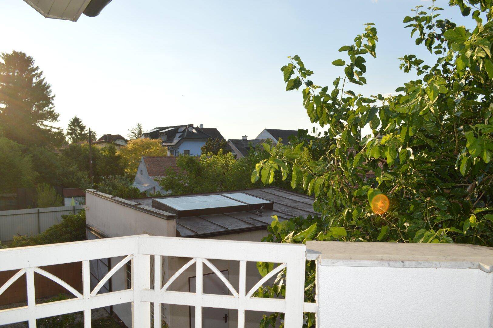 Blick vom Balkon auf die grüne Umgebung mit Bäumen und Dächern anderer Häuser in der Ferne.