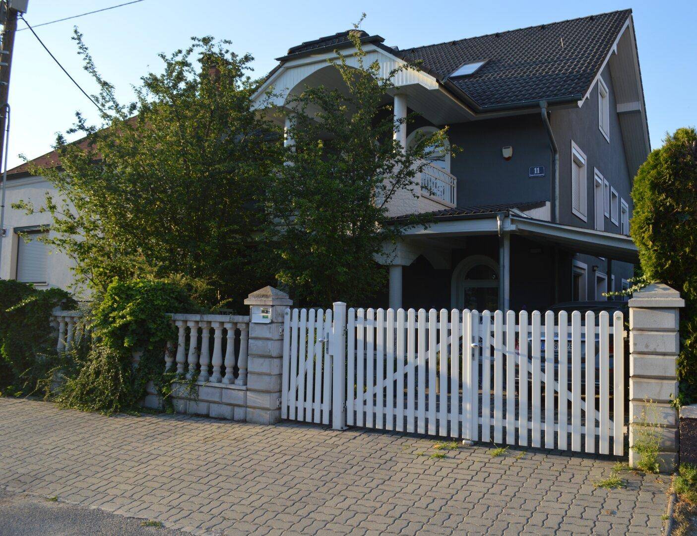 Das Haus präsentiert sich mit einer dunkelgrauen Fassade, einem weißen Zaun und üppigem Grün im Vorgarten.