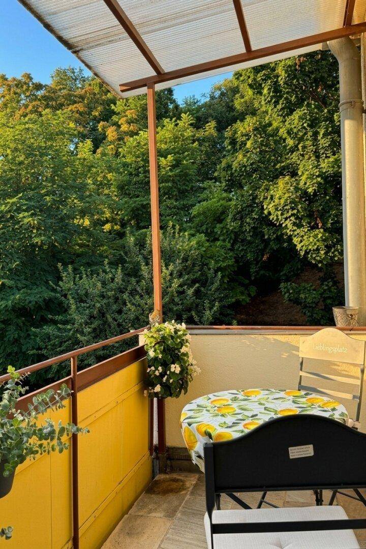 Sonniger Balkon mit Tisch und Stühlen, umgeben von üppigem Grün und Bäumen, ideal zum Entspannen.