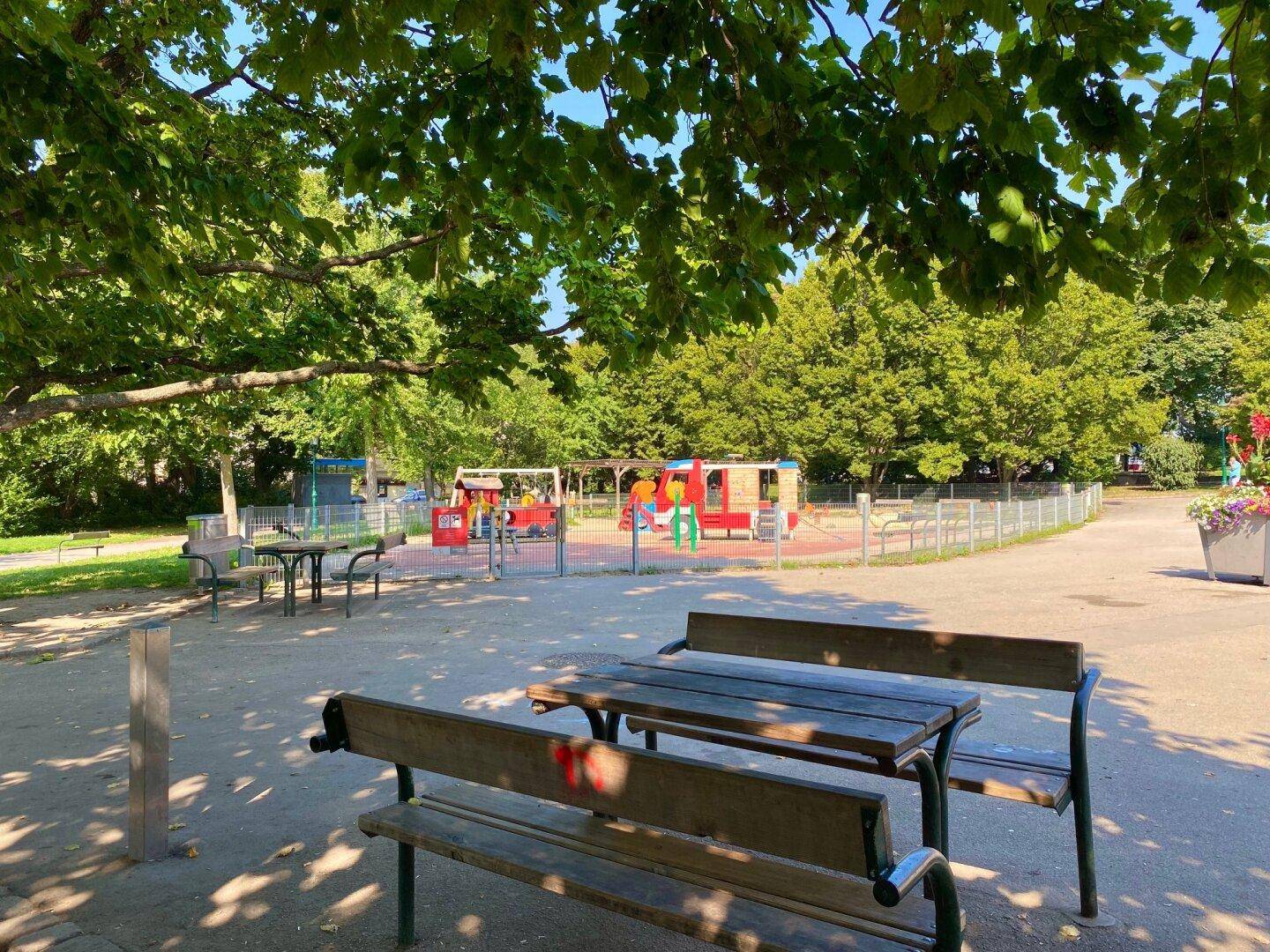 Sonniger Spielplatzbereich mit bunten Spielgeräten, umgeben von Bäumen und Sitzgelegenheiten.