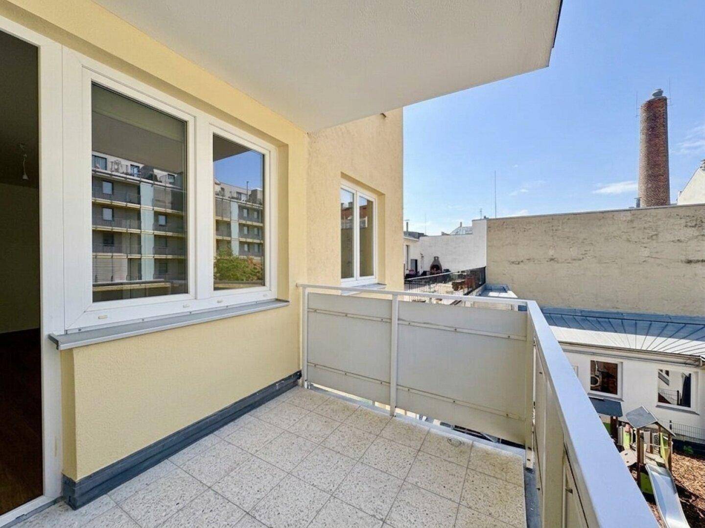 Kleiner Balkon mit Fliesenboden und Blick auf umliegende Gebäude und einen Spielplatz.