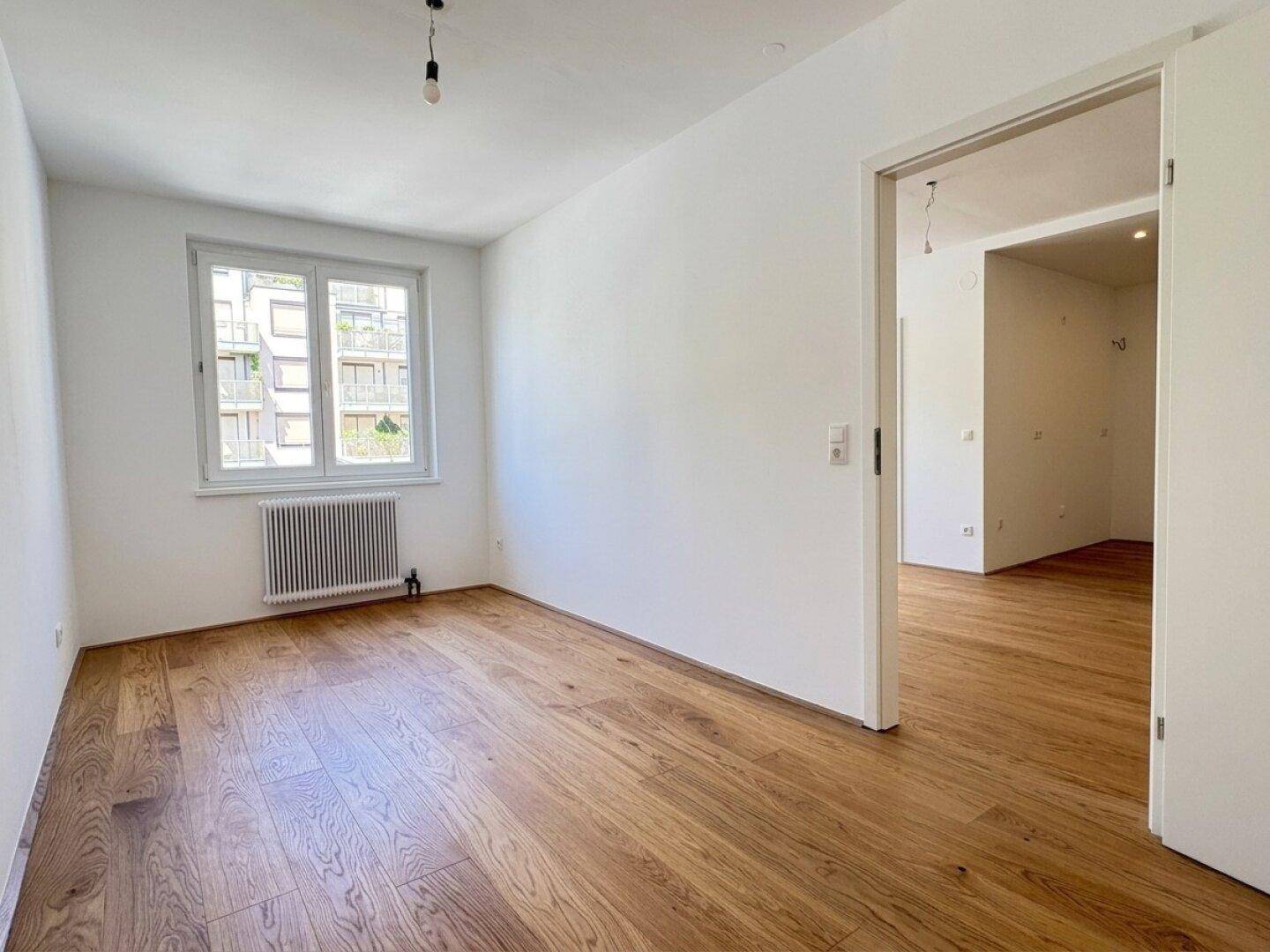 Helles Schlafzimmer mit Holzboden, Fenster und Blick auf die umliegenden Gebäude.