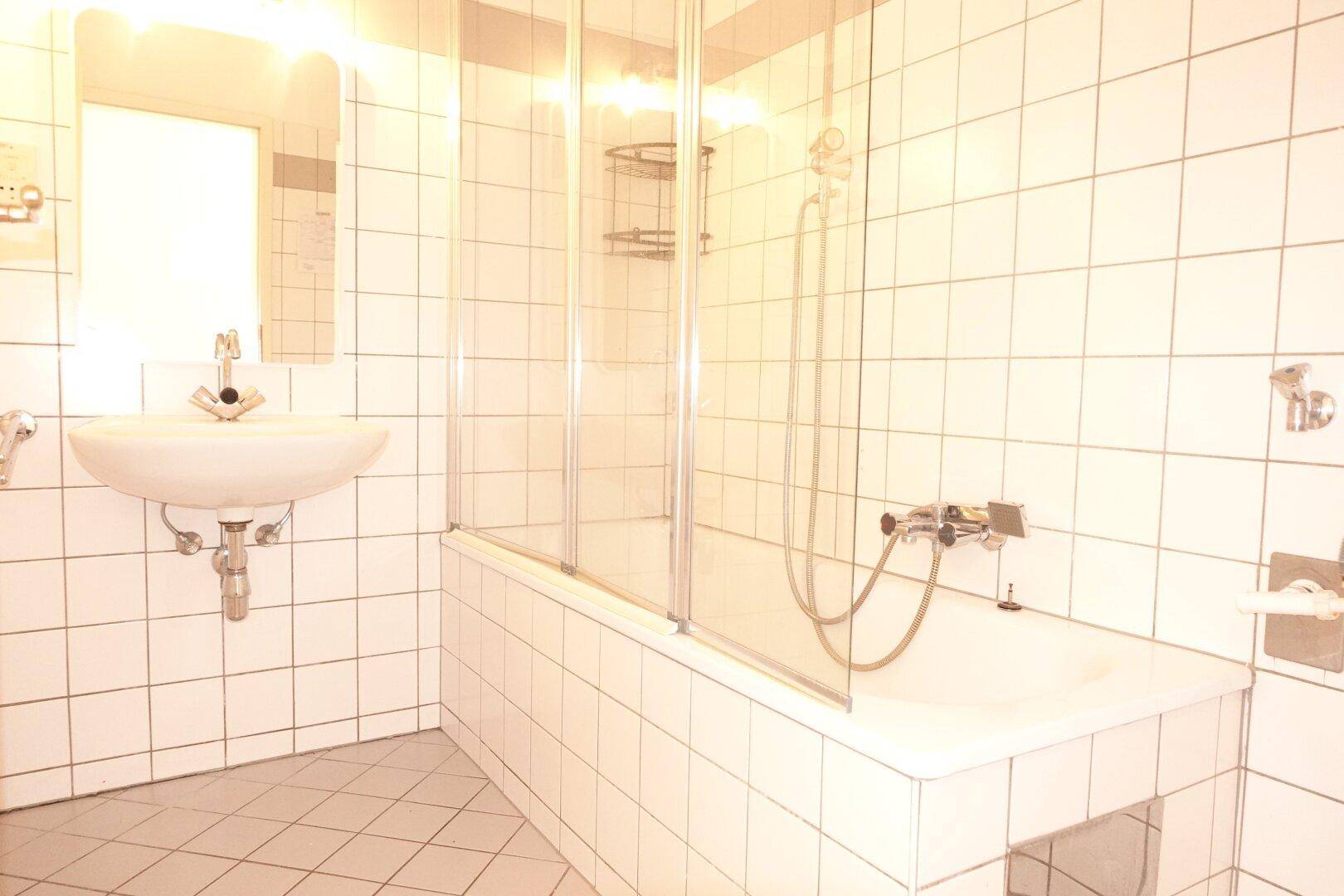 Helles Badezimmer mit weißen Fliesen, Waschbecken und Badewanne mit Duschabtrennung.