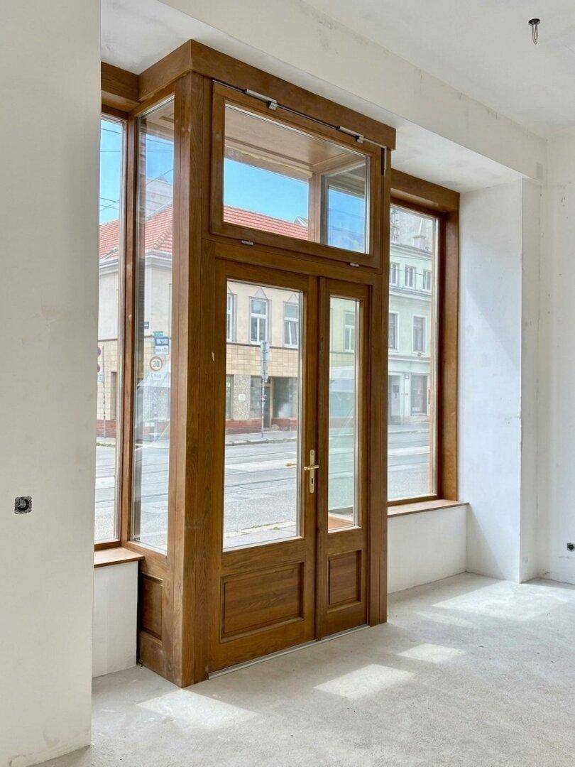 Innenansicht des eleganten Holzeingangs mit doppelten Glastüren und hohen Fenstern, die zur Straße zeigen.