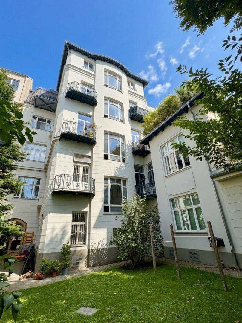 Mehrstöckiges Gebäude mit Balkonen, umgeben von einem grünen Garten unter blauem Himmel.