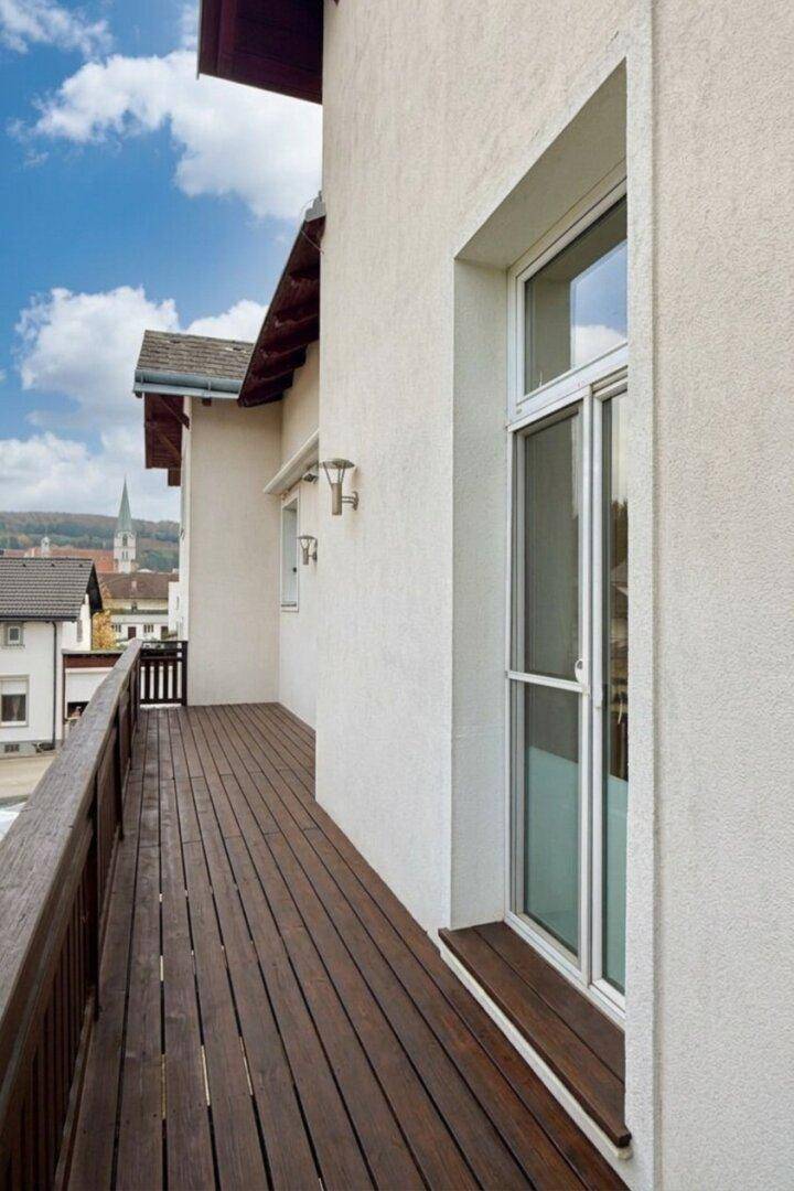 Langer Holzbalkon entlang der Hausfassade mit weißem Fenster und Blick auf die Nachbarschaft.