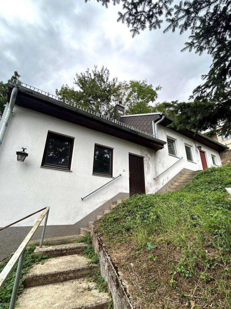 Außenansicht eines weißen Hauses an einem Hang mit Treppenaufgang unter bewölktem Himmel.