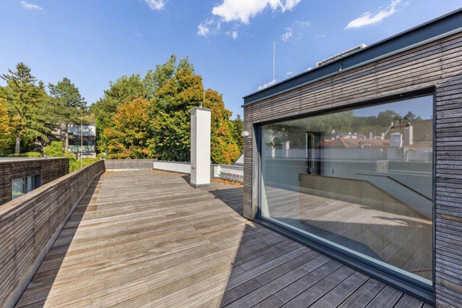 Moderne Dachterrasse mit Holzboden und großen Fenstern, umgeben von grünen Bäumen.