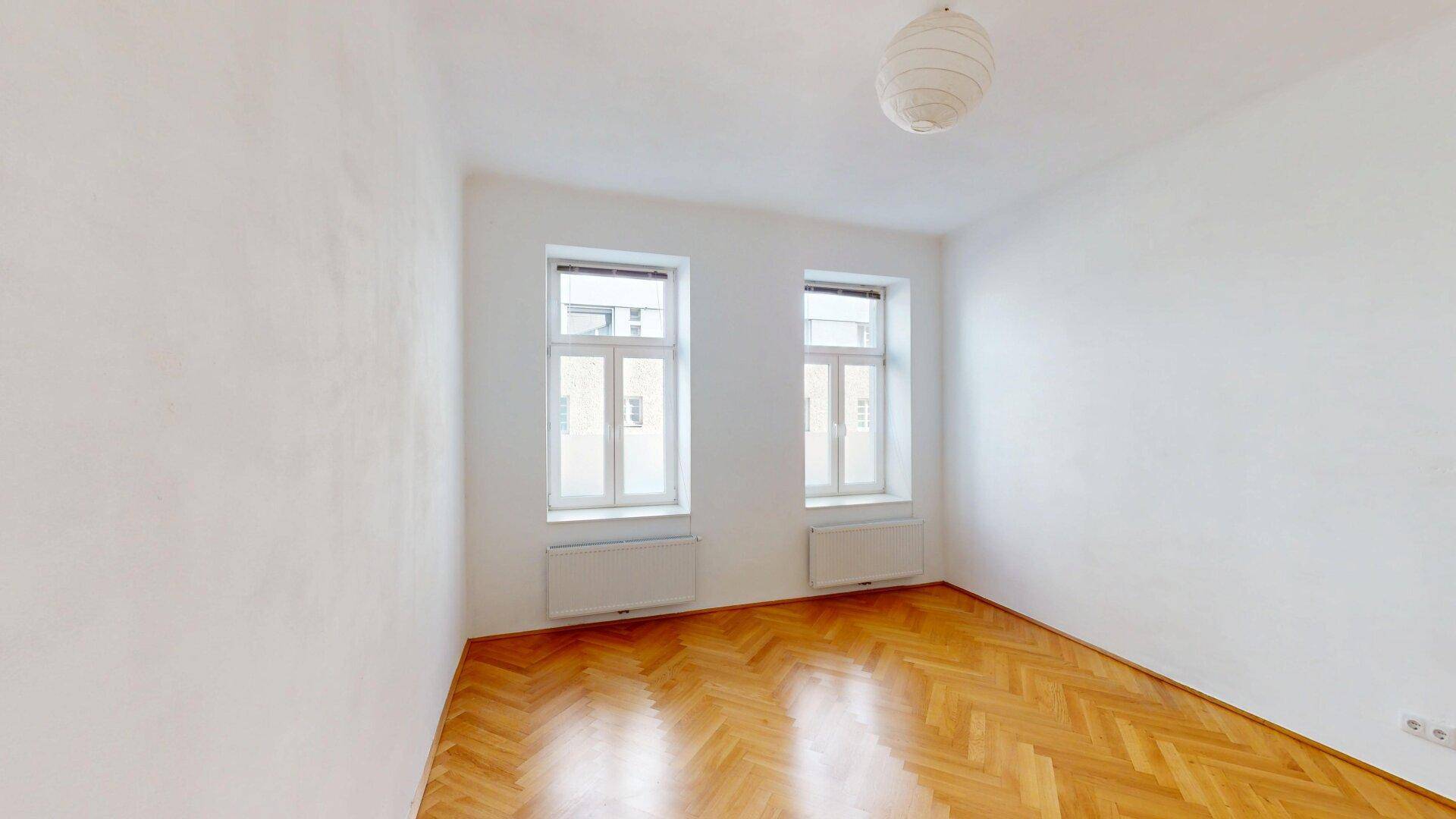 Lichtdurchflutetes Zimmer mit zwei Fenstern und glänzendem Fischgrätparkettboden.