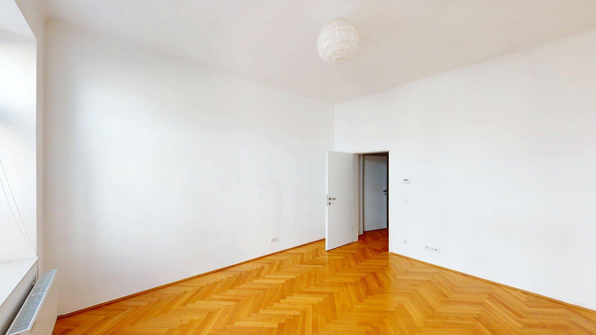 Geräumiges Zimmer mit hellem Fischgrätparkett und einer offenen Tür zu einem weiteren Bereich.