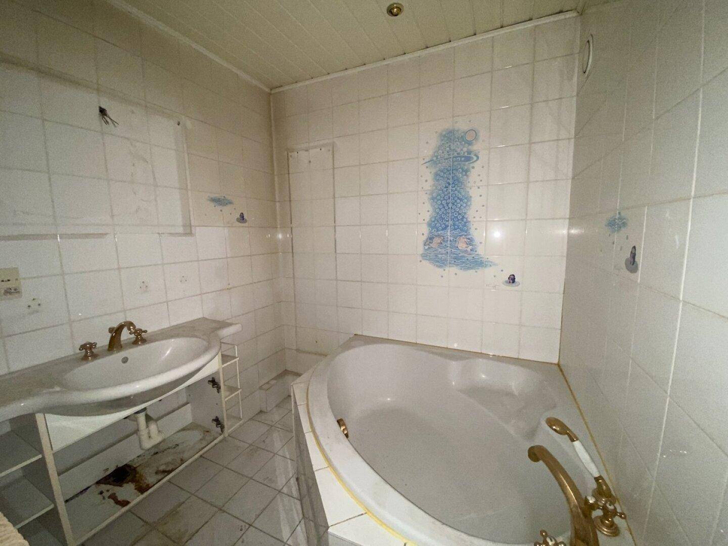 Veraltetes Badezimmer mit weißen Fliesen, Eckbadewanne und Einzelwaschbecken, renovierungsbedürftig.