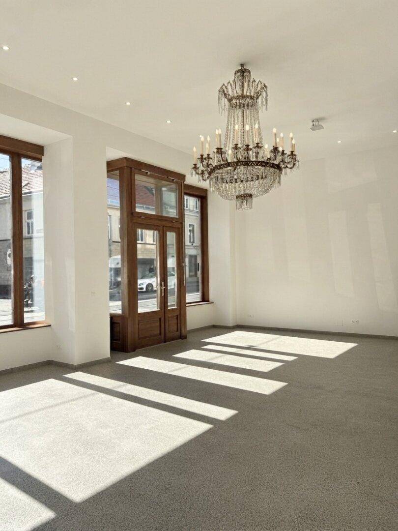 Großer, leerer Gewerberaum mit hohen Decken, elegantem Kronleuchter und breiten Fenstern zur Straße.