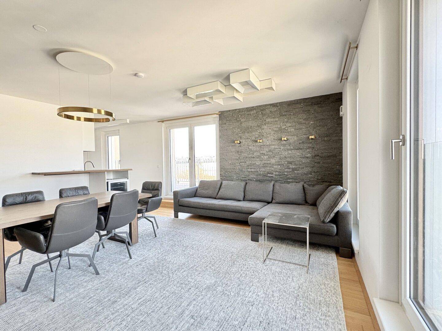 Offener Wohn-Essbereich mit großem Sofa, Esstisch und Zugang zu den Fenstern.