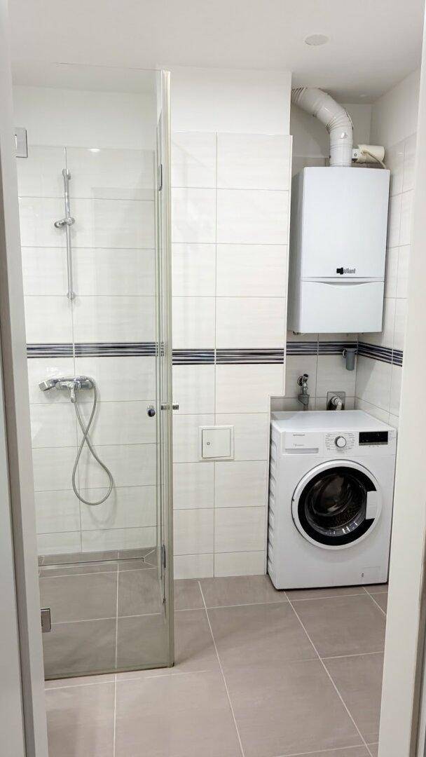 Modernes Badezimmer mit Duschkabine, Waschmaschine und Gastherme, hell gefliest.