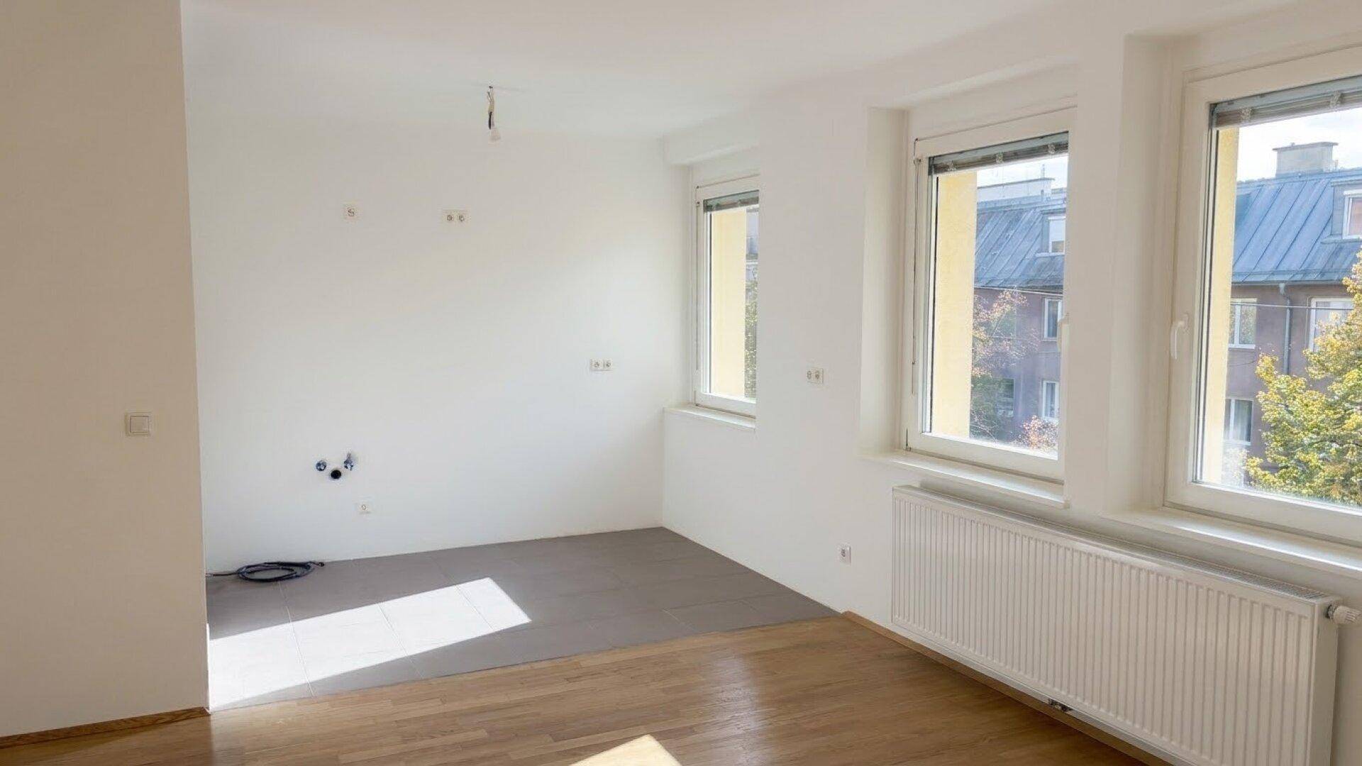 Heller Raum mit vorbereiteten Anschlüssen für eine Küche, kombiniert mit Parkett- und Fliesenboden.