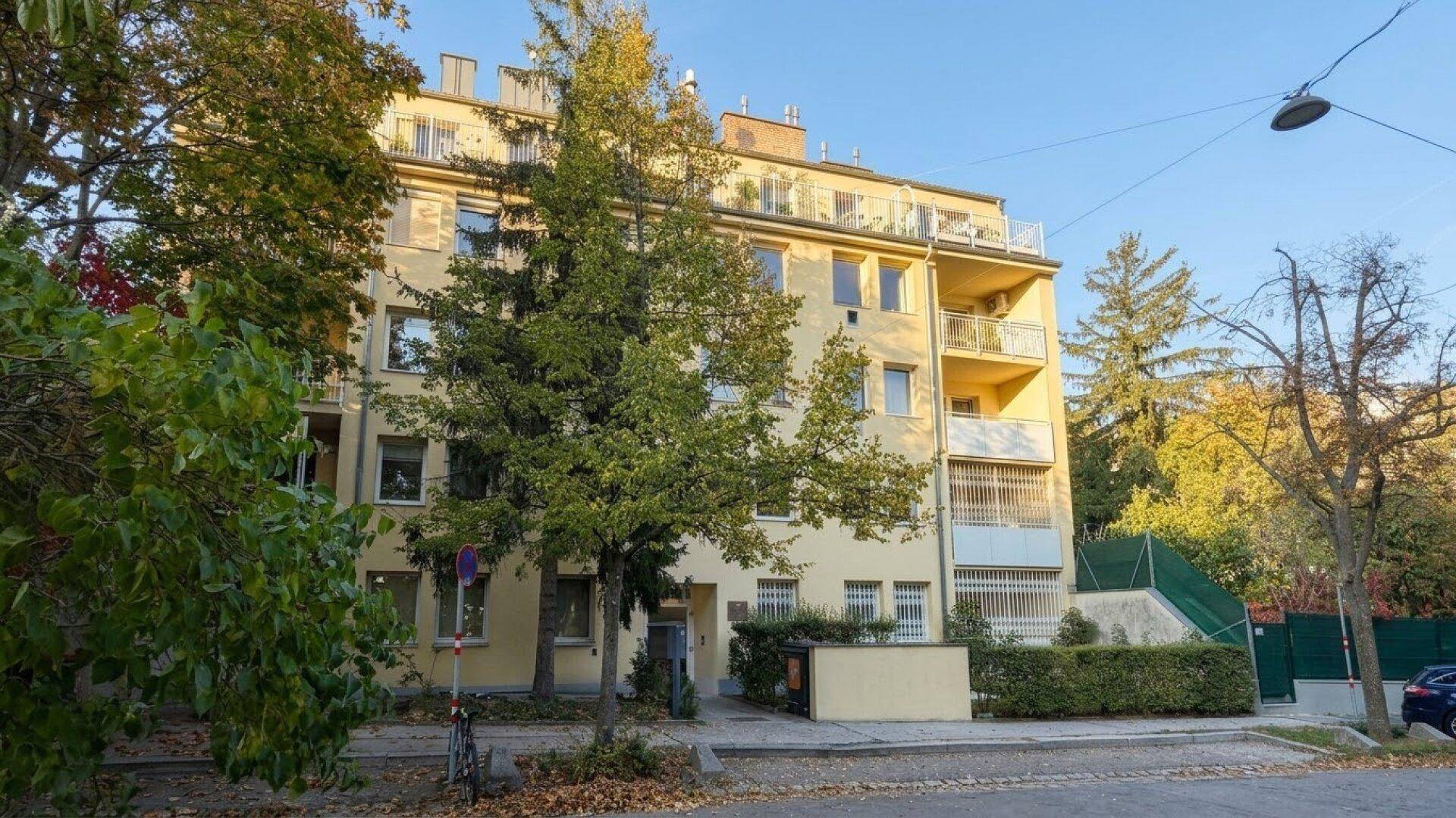 Gelbes Mehrfamilienhaus mit Balkonen und Bäumen im Vordergrund, an einem sonnigen Tag.
