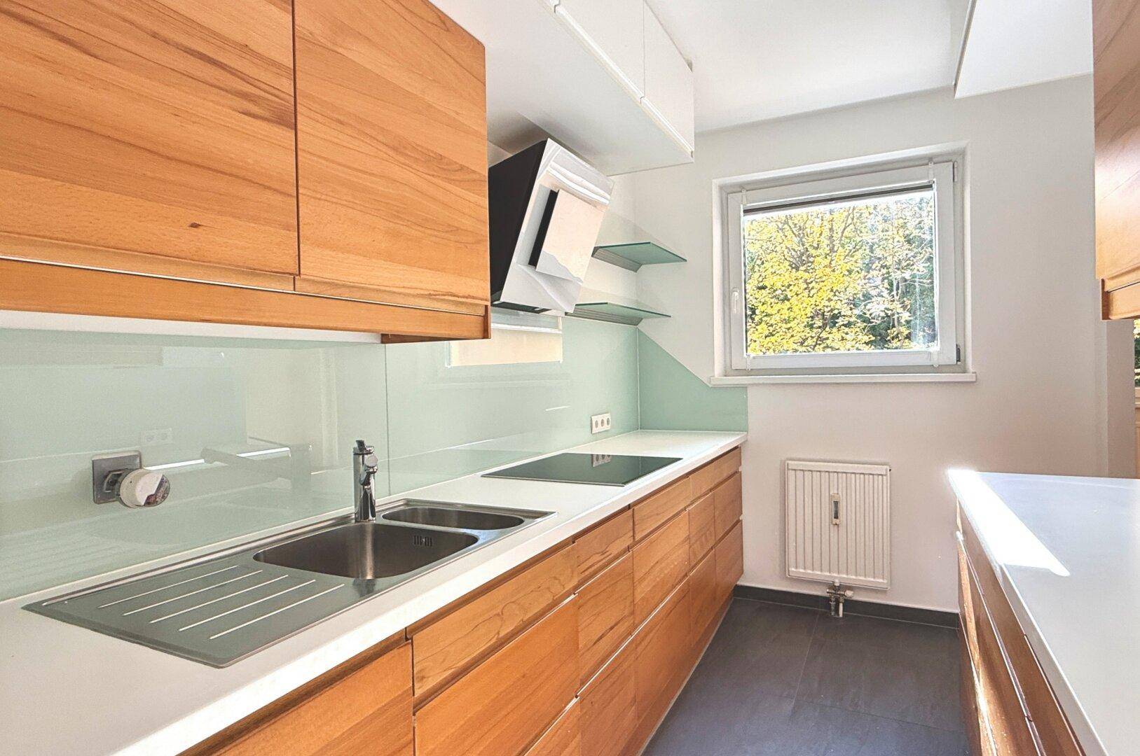 Moderne Küche mit hellen Holzfronten, weißer Arbeitsplatte und Fenster mit Grünblick.