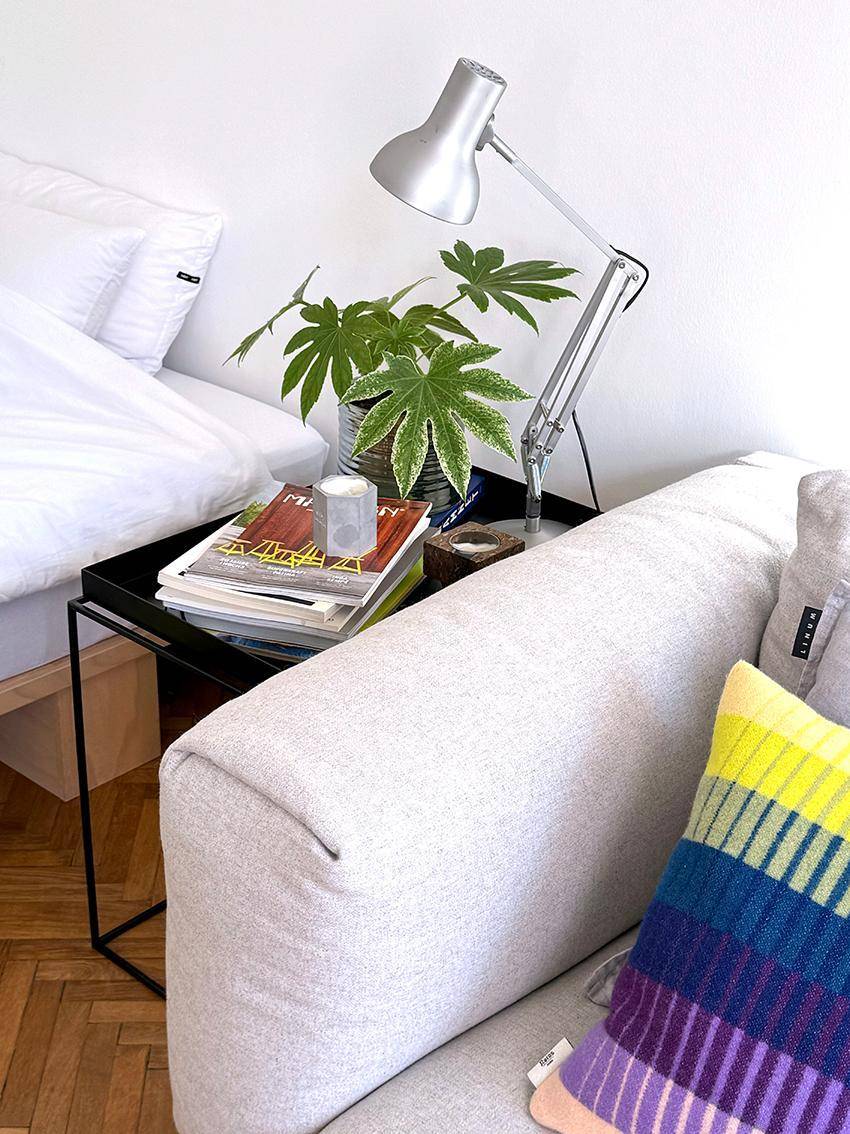 Nachttisch mit Lampe und Pflanzen zwischen Bett und Sofa im hellen Wohnbereich.