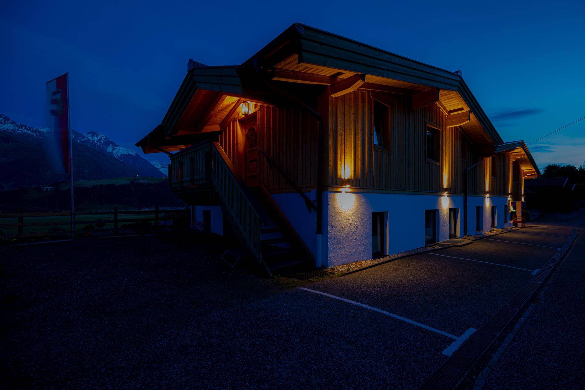 Das Gebäude ist nachts beleuchtet, mit sichtbaren Parkplätzen und einem Blick auf die Berge im Hintergrund.