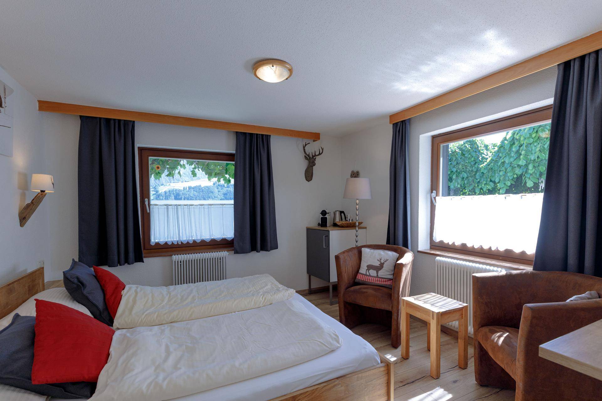 Helles Schlafzimmer mit Doppelbett, zwei Fenstern und rustikalen Holzmöbeln, die eine gemütliche Atmosphäre schaffen.