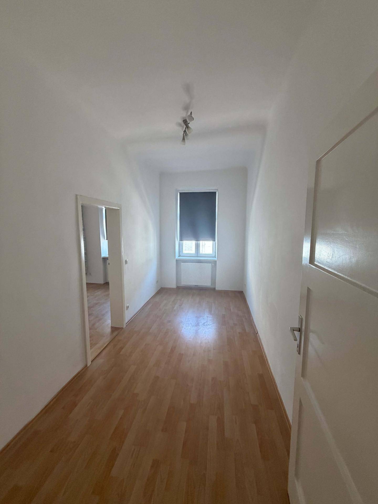 Langes, helles Zimmer mit Laminatboden und Fenster mit Rollo.