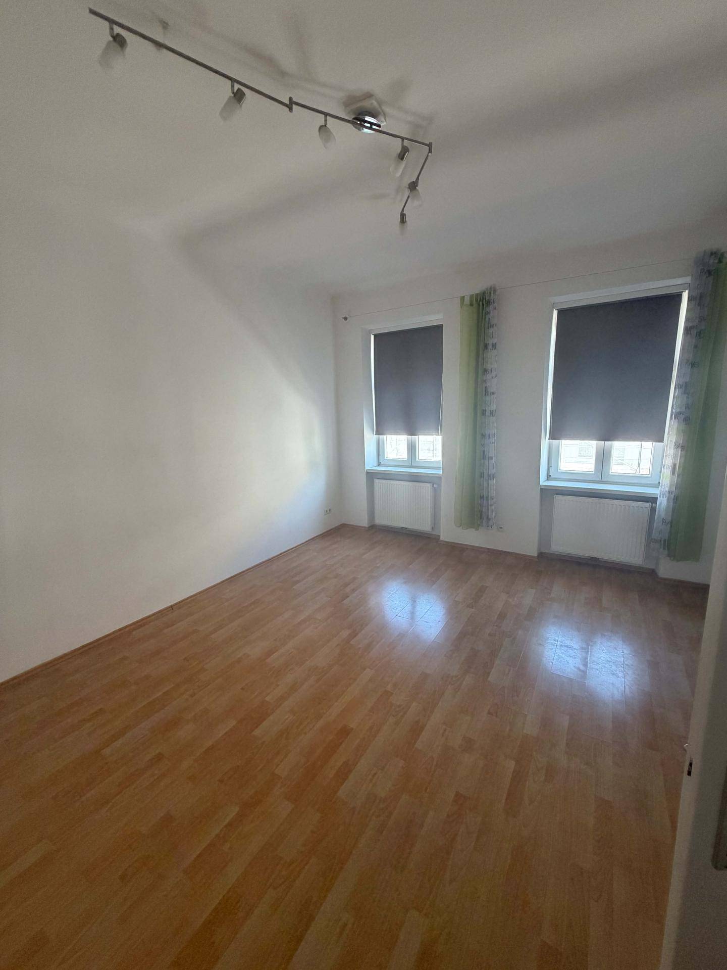 Helles Zimmer mit zwei Fenstern, Laminatboden und weißen Wänden.