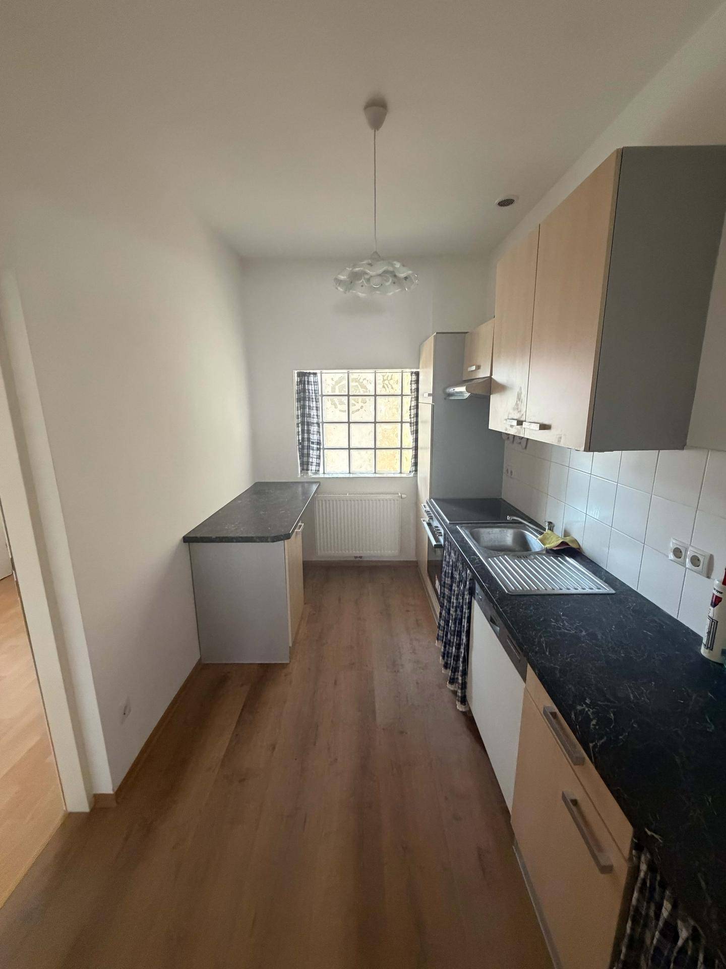 Einfache Küche mit Holzschränken, dunkler Arbeitsplatte und Fenster mit karierten Vorhängen.