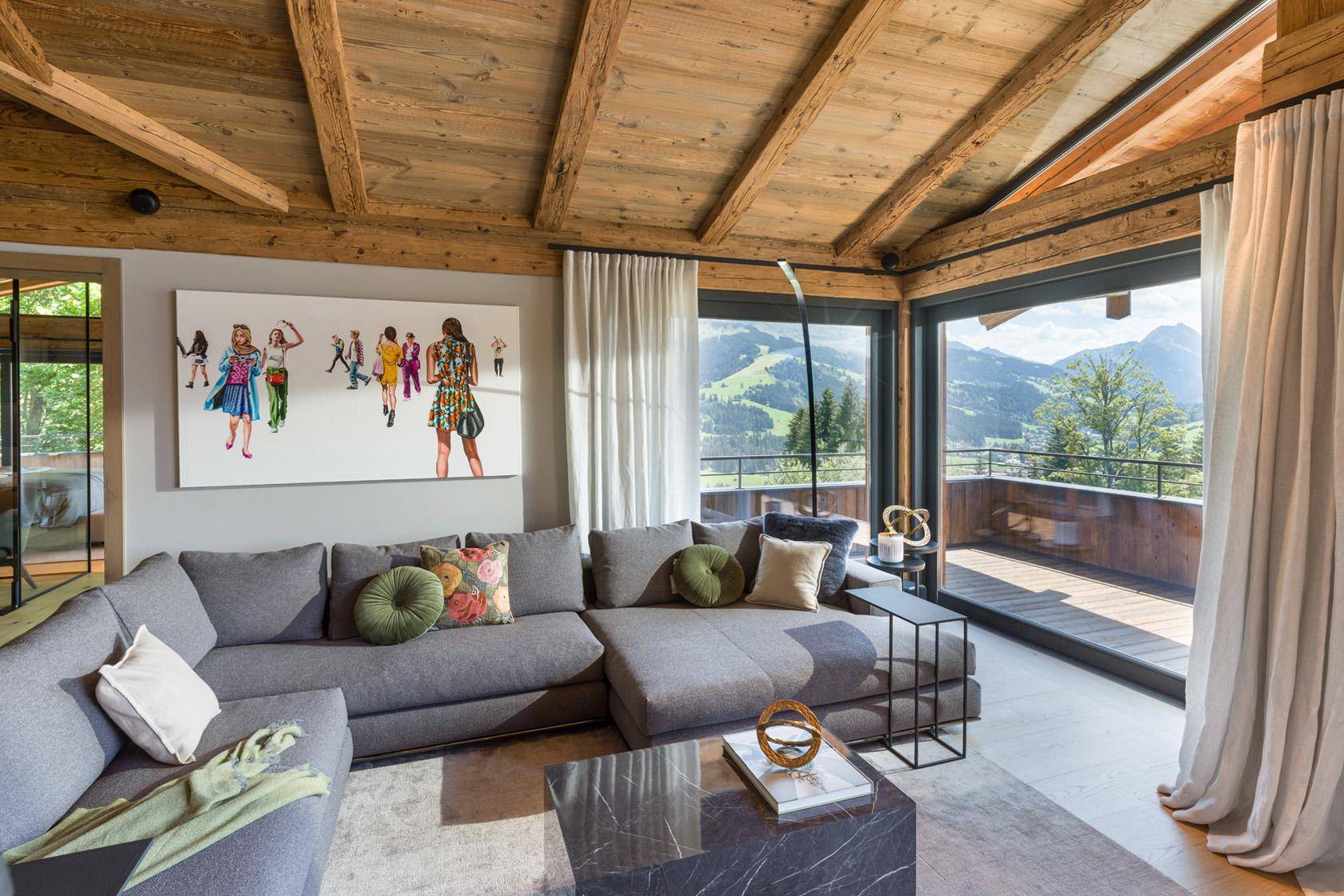 Gemütliches Wohnzimmer mit großem Sofa, Holzbalkendecke und Panoramablick auf die Berge.