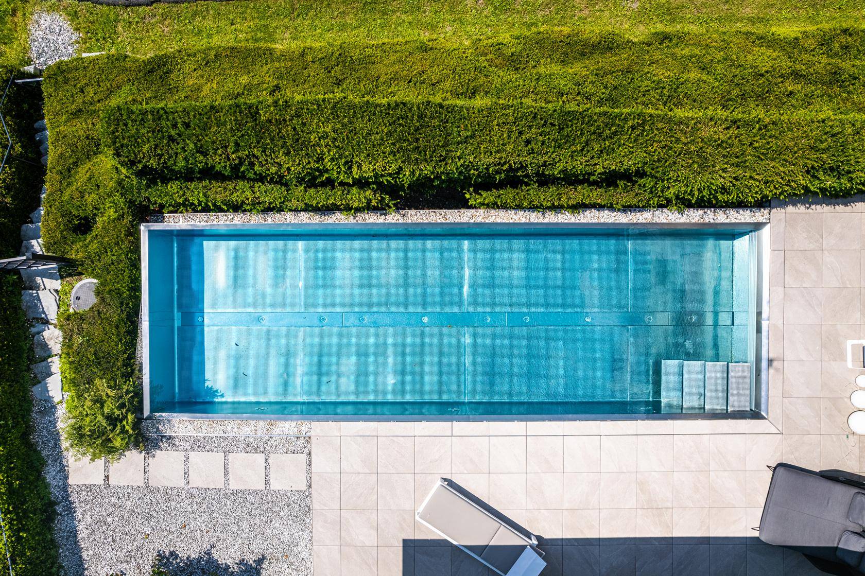 Vogelperspektive auf den rechteckigen Infinity-Pool mit umliegender Terrasse und Hecke.