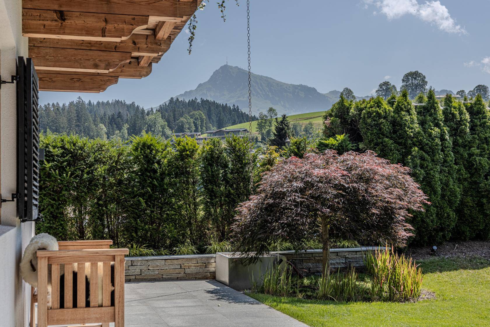 Sonniger Terrassenbereich mit Steinmauer, Zierbaum und Bergblick.