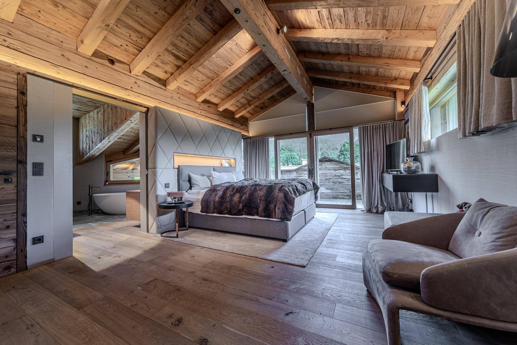 Gemütliches Schlafzimmer mit Holzbalkendecke, großem Bett und Zugang zu einem modernen Bad mit freistehender Wanne.
