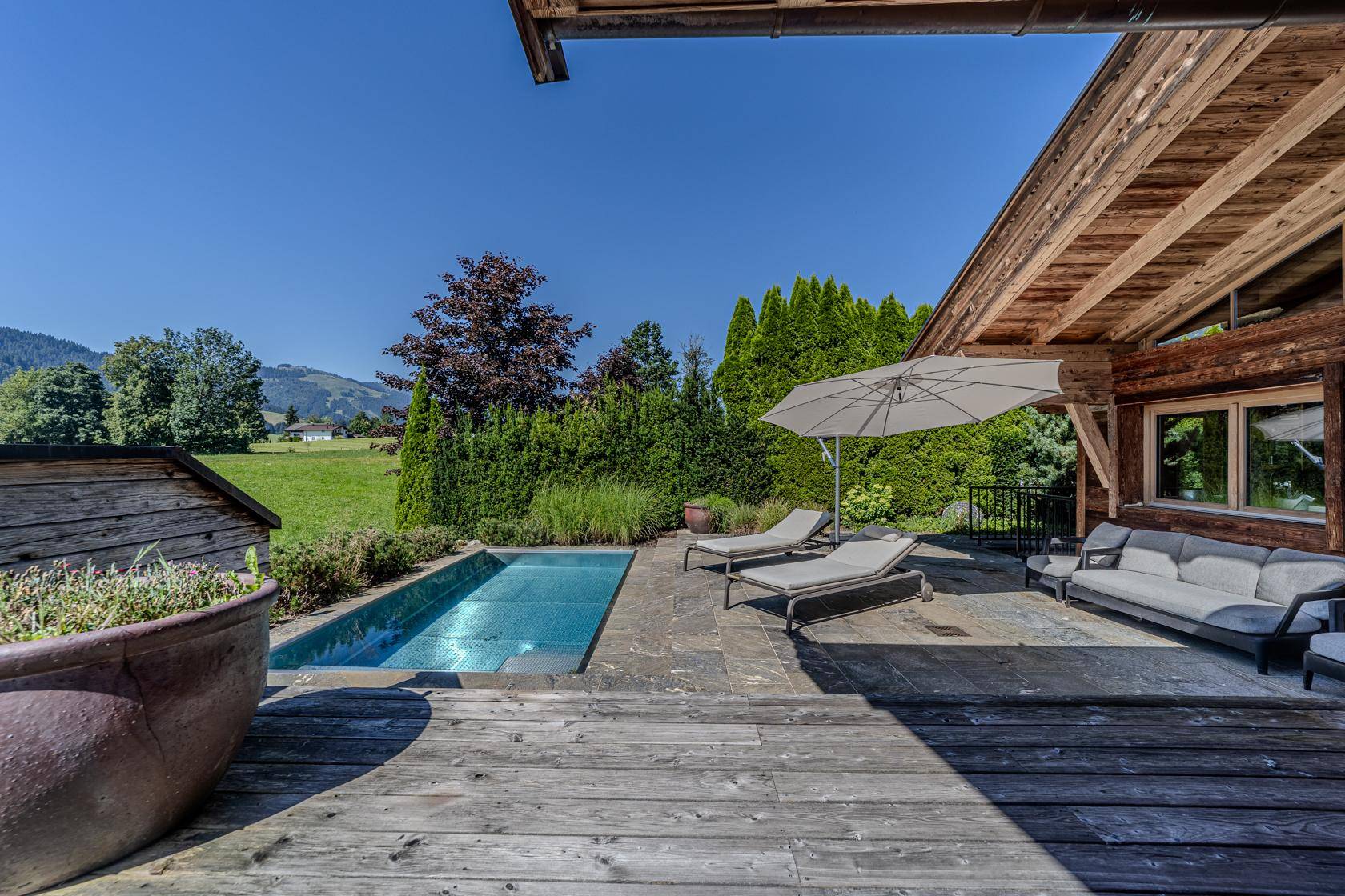 Sonniger Außenbereich mit kleinem Pool, Liegestühlen und weitem Blick auf die grüne Landschaft und die Berge.