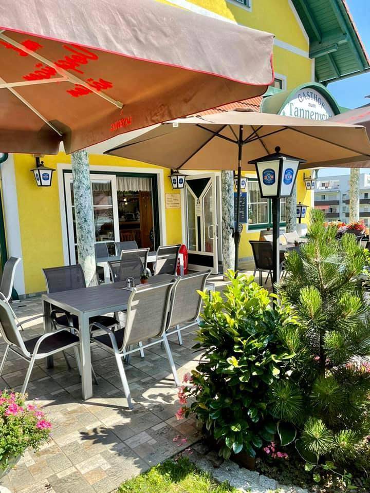 Helle Terrasse mit modernen Sitzmöbeln, Sonnenschirmen und dekorativen Pflanzen vor dem gelben Gebäude.