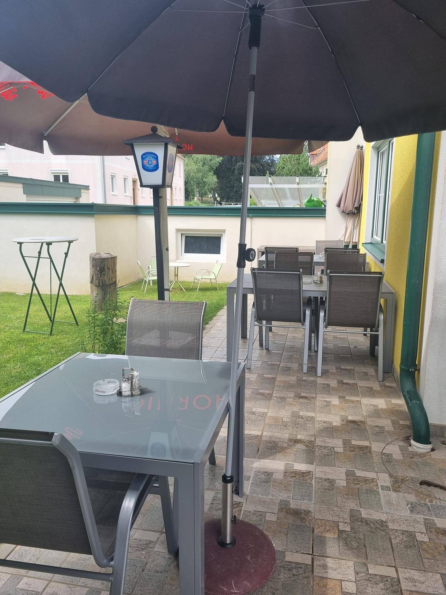 Gepflasterte Terrasse mit mehreren Tischen und Stühlen unter einem großen Sonnenschirm, angrenzend an eine Rasenfläche.