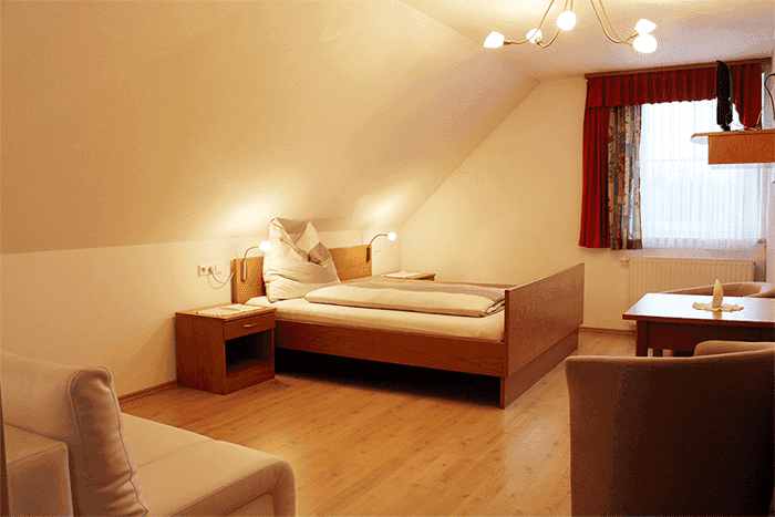 Helles Schlafzimmer im Dachgeschoss mit Holzbett, Sofa, Tisch und Laminatboden unter einer Dachschräge.
