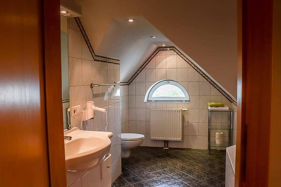 Helles Badezimmer mit Waschbecken, Toilette, Duschkabine und einem kleinen Rundfenster unter der Dachschräge.