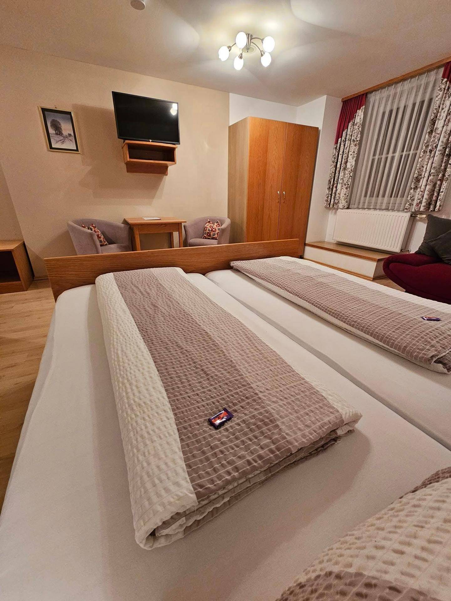 Schlafzimmer mit zwei Einzelbetten, Holzschrank, Fernseher und einem roten Sofa im Hintergrund.