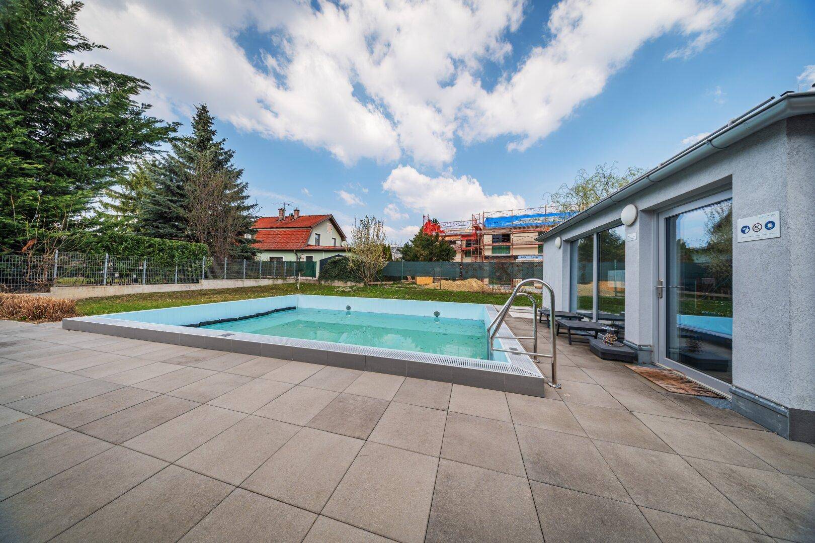 Sonniger Außenpool mit gepflasterter Terrasse und angrenzendem Gebäude, umgeben von Bäumen.